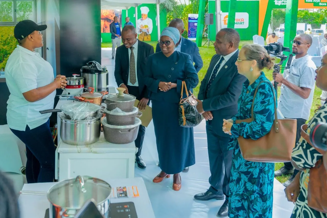 Officials and guests at a cooking demonstration stall, listening to an exhibitor beside pots and cookers